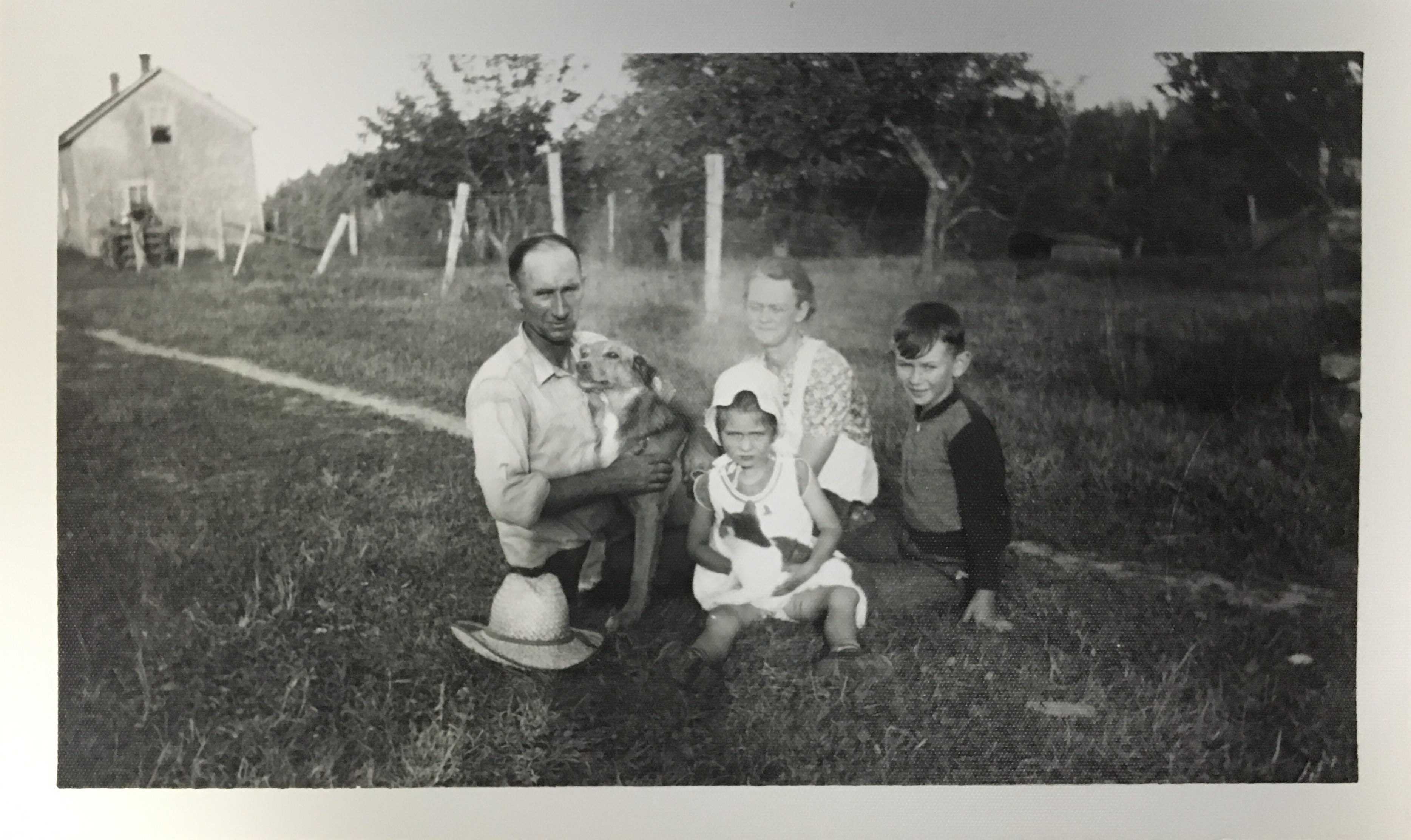 Photographie en noir et blanc – Archie et sa famille, assis sur la pelouse de leur ferme. Archie tient le chien de la famille, Brownie. Sa fille Margie, sa femme Grace et son fils Francis sont à sa gauche.